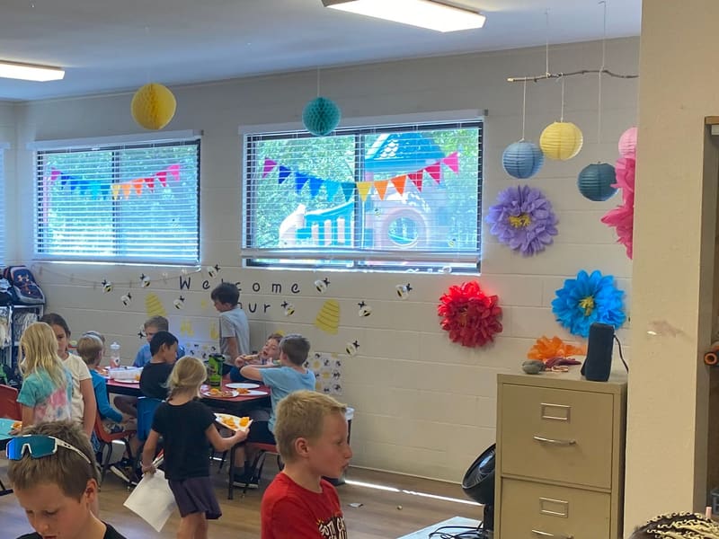 Campers at tables in the decorated camp room, with paper flowers and bunting on the windows