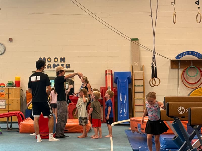 A Ninja Zone coach in a NINJA shirt with a group of kids in the gym, rings and beams nearby
