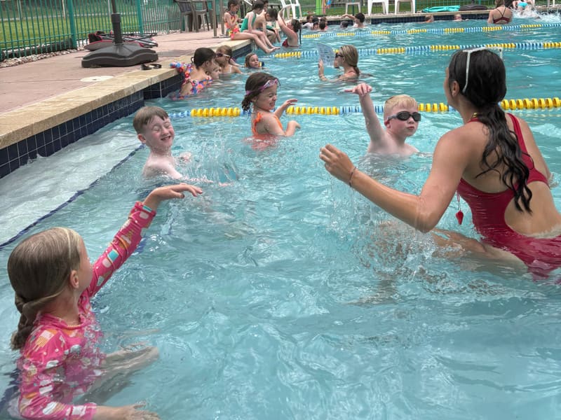 A swim instructor in a red suit splashing with smiling kids in the outdoor pool